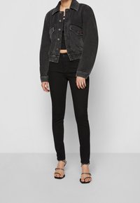 Chaqueta de mezclilla negra con un ligero desgaste, dos bolsillos y cierre de botones; combinada con jeans skinny negros y sandalias negras con tiras.