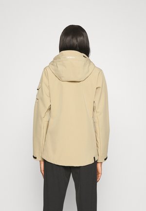 Personne portant une veste beige à capuche et un pantalon noir, vue de dos sur un fond clair uni.