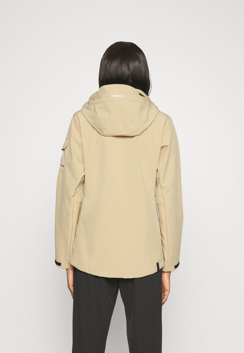 Personne portant une veste beige à capuche et un pantalon noir, vue de dos sur un fond clair uni.