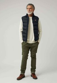 Chaleco de plumas azul marino sobre un suéter crema, pantalones verde oliva y botas marrones con cordones. El chaleco tiene un cuello alto y cierre de cremallera.