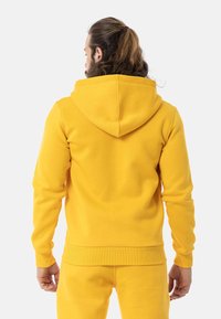 Gelbes Kapuzen-Sweatshirt mit lockerer Passform, gerippten Bündchen und Saum. Weicher Stoff, Kängurutasche und Nahtdetails an der Kapuze.