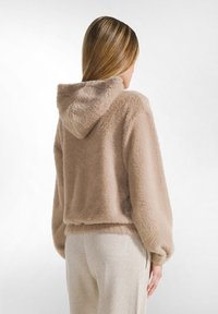 Sudadera beige, esponjosa, con un acabado texturizado, que presenta una tela suave, un corte relajado y capucha. Combinada con pantalones de color claro y brillantes.