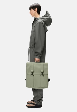 Mochila estructurada verde con dos correas frontales, combinada con una chaqueta gris, pantalones grises y chanclas negras. Diseño plano y rectangular.