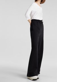 Zwarte, brede broek met een elastische tailleband en verstelbaar trekkoord. Gladde stof; gecombineerd met een wit, lang mouwtop en sneakers.