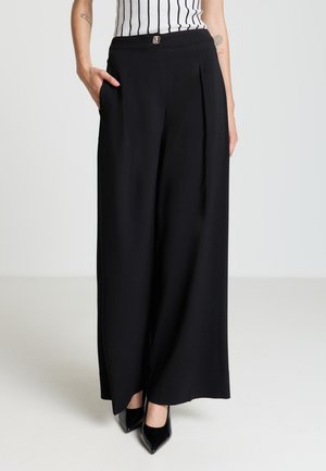 Pantaloni neri a gamba larga con pieghe, realizzati in tessuto leggero. Dotati di tasche frontali e chiusura con bottone. Indossati con tacchi neri a punta.