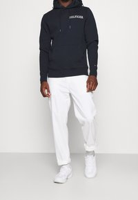 Sudadera marina con logo blanco, bolsillo delantero, capucha con cordón, combinada con pantalones blancos y zapatillas ligeras. Conjunto sencillo y casual.