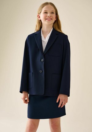 Lächelndes Mädchen, das einen marineblauen Blazer mit Knöpfen und Taschen über einem weißen Hemd und einem passenden marineblauen Rock trägt, steht vor einem einfarbigen Hintergrund.