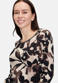 T-shirt à manches longues en beige avec des motifs floraux sombres. Bordure du col noire. Tissu doux et texturé, design ajusté et ourlet arrondi.