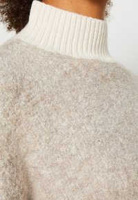 Hellbeige gestrickter Pullover mit einem gerippten weißen Rollkragen. Die Textur ist weich und flauschig, mit einer gleichmäßigen Dichte im gesamten Stoff.