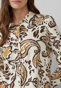 Vrouw in een lange mouwen overhemd met een beige, bruine en mosterdkleurige bloemen- en paisleypatroon, kraag zichtbaar.