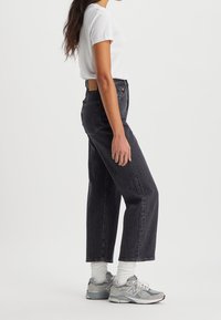 Jean en denim noir taille haute avec une coupe décontractée, tissu texturé, et patch en cuir à l'arrière. Associé à un T-shirt blanc et des baskets grises.