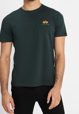 Homme portant un t-shirt Alpha Industries vert foncé avec un petit logo jaune sur la poitrine, associé à un pantalon noir.