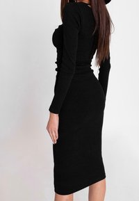 Donna con capelli lunghi che indossa un vestito midi nero a coste, aderente e con maniche lunghe, di profilo contro uno sfondo neutro.