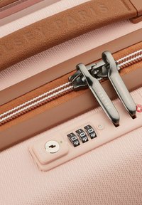Valise rose avec un extérieur texturé, une fermeture éclair accentuée de marron, deux fermetures éclair argentées et un verrou à combinaison avec un trou de clé.