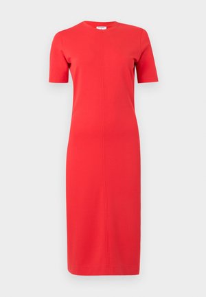 Kvinde med mørkt hår iført en tætsiddende rød knælængde kjole, der holder en sort håndtaske i venstre hånd, iført sorte flade sko, stående mod en hvid baggrund.