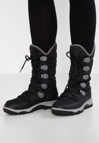 Bottes isolantes noires avec une texture matelassée, des accents gris et un laçage à l'avant. Elles présentent une semelle épaisse et robuste pour une meilleure traction.