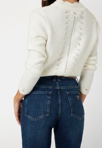Weißer Strickpullover mit einem Spitzenmuster auf der Rückseite, kombiniert mit dunklen Jeans aus Denim, die über Gesäßtaschen und ein Logo-Detail verfügen.