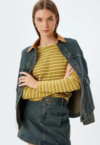 Jeansjacke mit einem hellbraunen Cordkragen, über ein gelb gestreiftes Langarmshirt und einen Jeansrock getragen. Minimalistisches Design, lässige Passform.