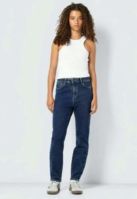 Modri jeansi iz denima z visokim pasom, ravnim krojem in klasičnim petim žepnim dizajnom, v kombinaciji z belo majico brez rokavov in svetlimi supergami.