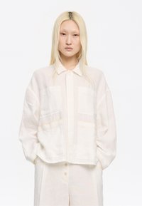Cropped light beige shirt with a button-up front, two front pockets, and subtle stitching details. Made from a textured linen fabric.