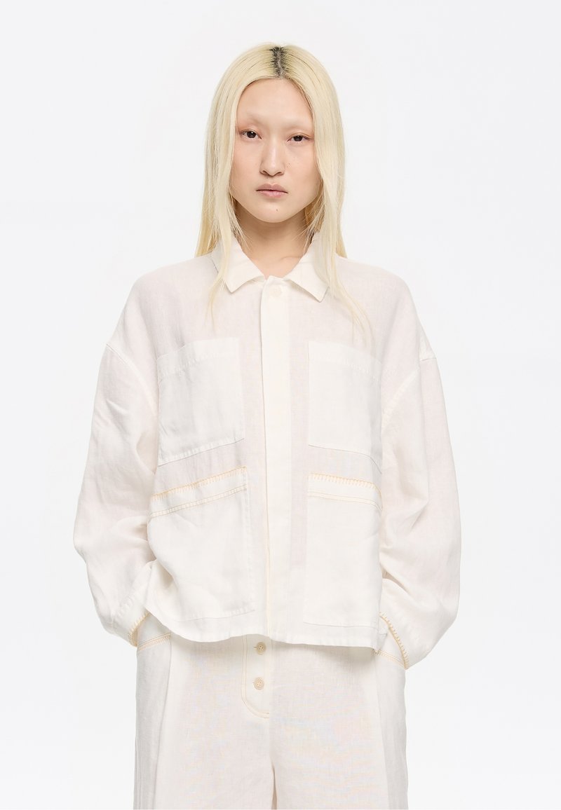 Geknipte lichtbeige blouse met een knoopsluiting aan de voorkant, twee voorzakken en subtiele stikdetails. Gemaakt van een gestructureerde linnen stof.