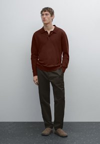Polo shirt in maglia marrone con colletto rilassato, abbinato a pantaloni scuri e scarpe slip-on marroni, su uno sfondo neutro.