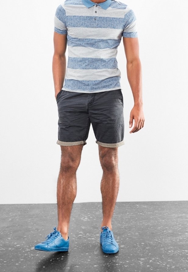 Homme debout portant un polo rayé bleu et blanc, un short gris foncé retroussé et des baskets bleu vif sur un sol moucheté.