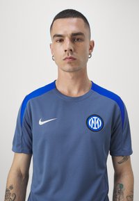 Camiseta atlética gris con acentos azules, que presenta un logo redondo y un swoosh de Nike. Hecha de un tejido texturizado y transpirable.