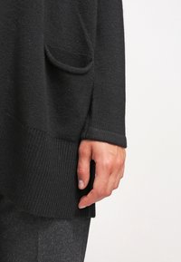 Main reposant à côté de la poche d'un pull en maille foncé, portant un pantalon gris foncé sur un fond clair uni.