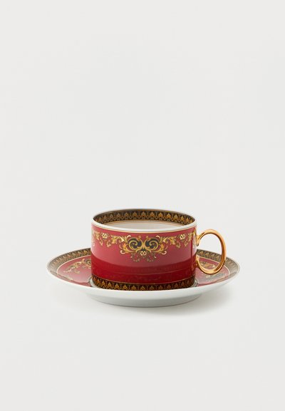 Tasse à café rouge et soucoupe avec motifs floraux dorés et une anse dorée, remplie d'une boisson brun clair, sur un fond blanc.
