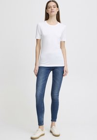 Bílá krátká tričko s krátkým rukávem v kombinaci s modrými skinny džíny a bílými teniskami s béžovou podrážkou, zobrazené na stojícím modelu na neutrálním pozadí.