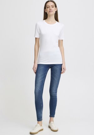 Hvit t-skjorte med kort erm kombinert med blå skinny jeans og hvite sneakers med beige såle, vist på en stående modell med en nøytral bakgrunn.