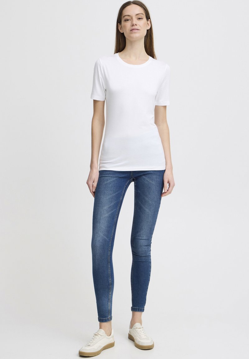 Bílá krátká tričko s krátkým rukávem v kombinaci s modrými skinny džíny a bílými teniskami s béžovou podrážkou, zobrazené na stojícím modelu na neutrálním pozadí.