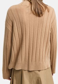 Una donna che indossa un maglione a coste beige e una gonna plissettata beige abbinata, vista da dietro su uno sfondo bianco.