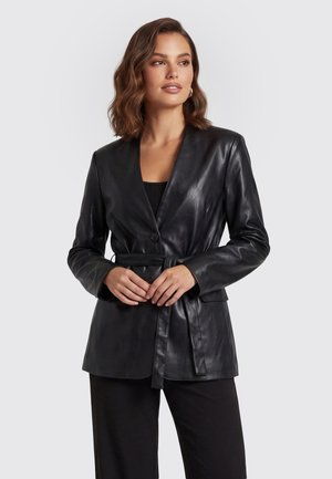 Vrouw draagt een zwarte leren blazer met riem over een zwarte broek, staand voor een eenvoudige lichte achtergrond, handen in de taille gevouwen.
