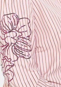 Bledoružová pruhovaná blúzka so vzorom kvetov vytvoreným z flitrov v odtieňoch ružovej a čiernej na ľavom rukáve, textúrovaná látka.