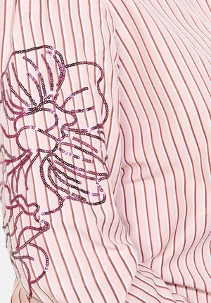 Blouse rayée rose clair avec un design floral en sequins dans des nuances de rose et de noir sur la manche gauche, tissu texturé.