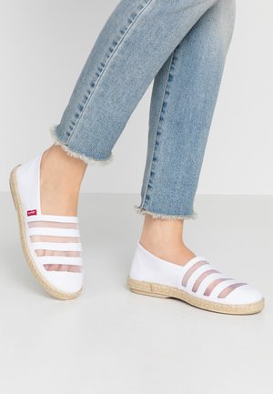Jambes portant un jean bleu clair à ourlet effiloché et des chaussures slip-on blanches avec des bandes transparentes en mesh horizontal et des semelles beige tissées.