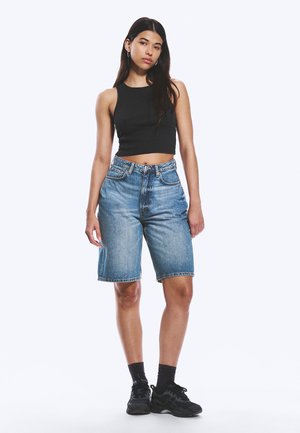 Ung kvinne står iført en svart crop top, høytlivs denimshorts til knelengde, svarte sokker og svarte joggesko.