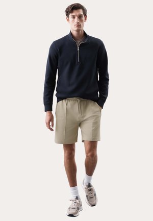 Mand iført en sort pullover med kvart-lynlås, beige shorts med snøre, hvide sokker og beige sportssko, stående foran en ensfarvet baggrund.