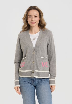 Frau trägt einen grauen, geknöpften Cardigan mit pinken Buchstaben "LO" und "VE" auf den Taschen, darunter ein weißes Hemd und blaue Jeans, steht vor weißem Hintergrund.