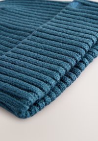 Tirkizno rebrasta odeja z mehko teksturo, z dvignjenimi črtami in robom z naborkom. Vsebuje majhno znamkarsko etiketo.