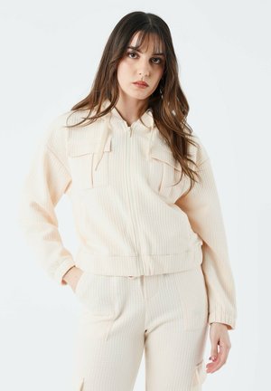 Helle Pfirsich-Fleecejacke mit strukturiertem Stoff, zwei vorderen Taschen und gerippten Bündchen. Kombiniert mit passenden strukturierten Hosen.