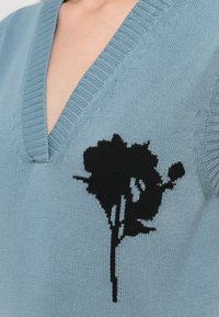 Lys blå strikkegenser med dyp V-hals og et svart blomsterdesign på forsiden, som fremhever tekstur og designdetaljer.