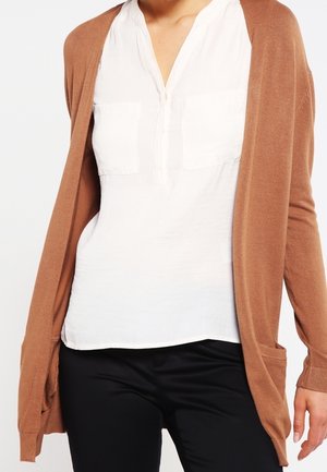 Femme portant un chemisier blanc, un pantalon noir et un cardigan marron long à manches longues et ouvert, avec des poches à l’avant.