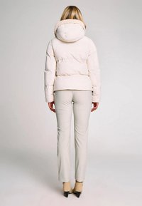 Donna con giacca imbottita beige chiaro con cappuccio, pantaloni beige aderenti e stivaletti con tacco beige, in piedi di spalle su uno sfondo neutro.