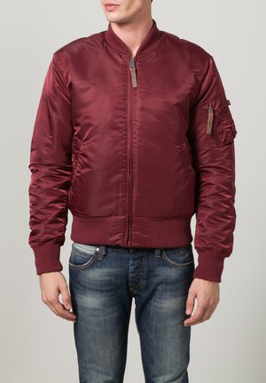 Mann, der eine burgunderfarbene Bomberjacke mit Reißverschluss, gerippten Bündchen und Saum trägt, kombiniert mit abgewetzten blauen Jeans vor einem neutralen Hintergrund.