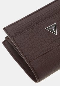 Portafoglio in pelle textured marrone scuro con cuciture visibili e un piccolo logo triangolare in metallo Guess USA sul fronte.