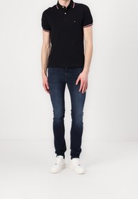 Muž v černém polo tričku, tmavě modrých slim fit džínách a bílých teniskách, stojící proti čistému bílému pozadí.