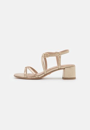 Sandale dorée à brides avec un petit talon bloc et une boucle ajustable à la cheville sur un fond clair uni.